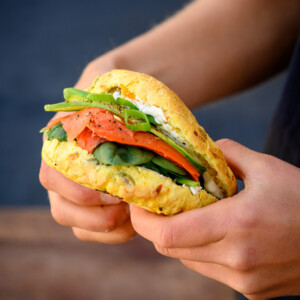 A photo of a Bodhi's Pumpkin & Turmeric Spelt Sourdough roll filled with Ricotta Cheese, 2 Slices Smoked Salmon, 1 Spring onion, sliced and 1/2 cup baby spinach leaves held between two hands